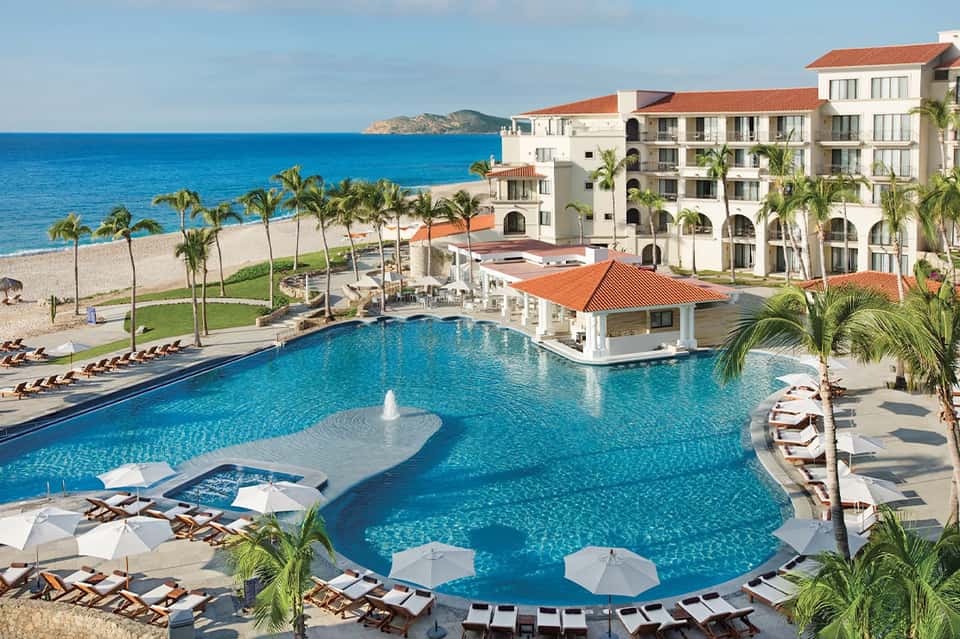 Beachfront resort pool with palm trees, lounge chairs, umbrellas, and ocean views