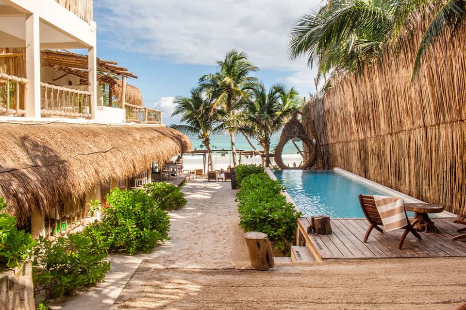 Beachfront resort pathway with thatched-roof buildings, palm trees, and blue pool with ocean beyond