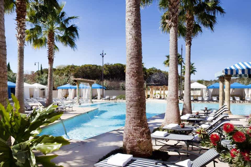 Large swimming pool with tall palm trees, lounge chairs, and covered cabanas