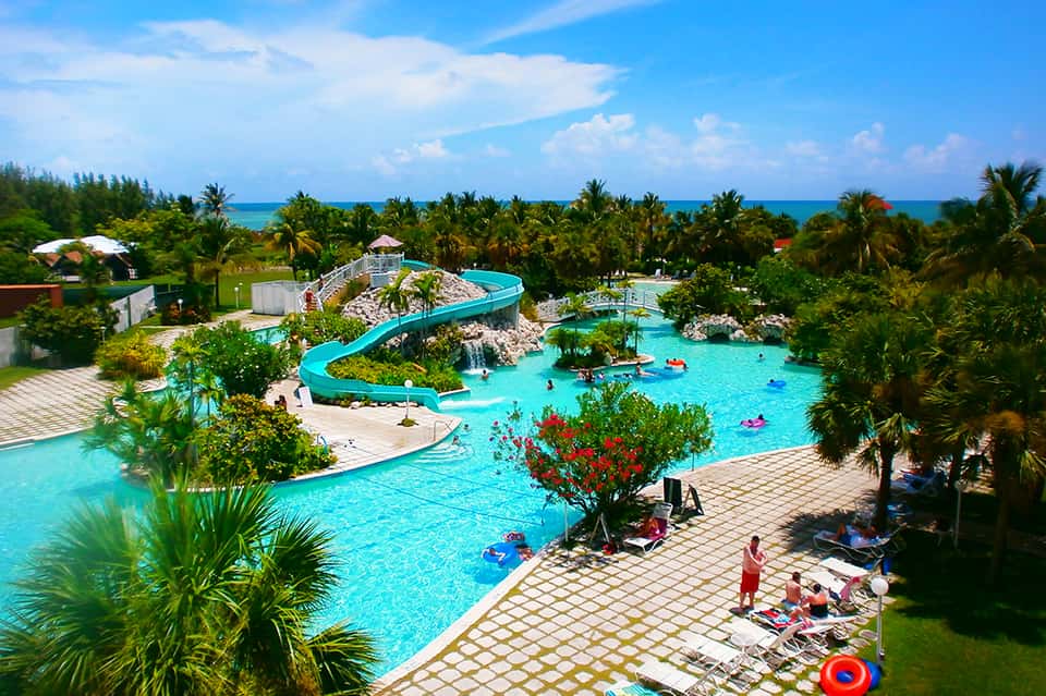 Resort water park with turquoise pools, blue water slide, palm trees, and people swimming