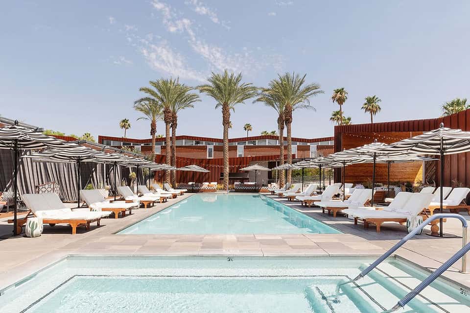 Resort pool with palm trees, white loungers, striped umbrellas, and rust-colored building