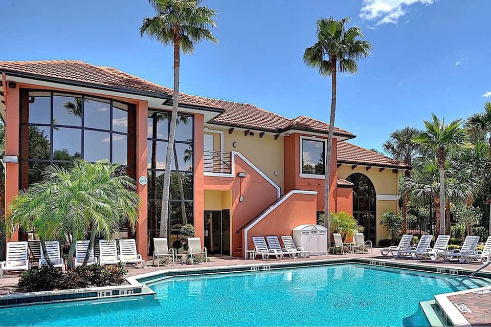 Resort pool area with coral-colored buildings, palm trees, white loungers, and clear blue sky