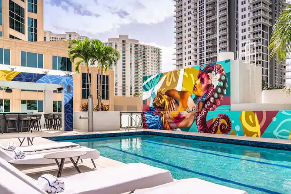 Urban rooftop pool with colorful pop-art mural, lounge chairs, and city buildings