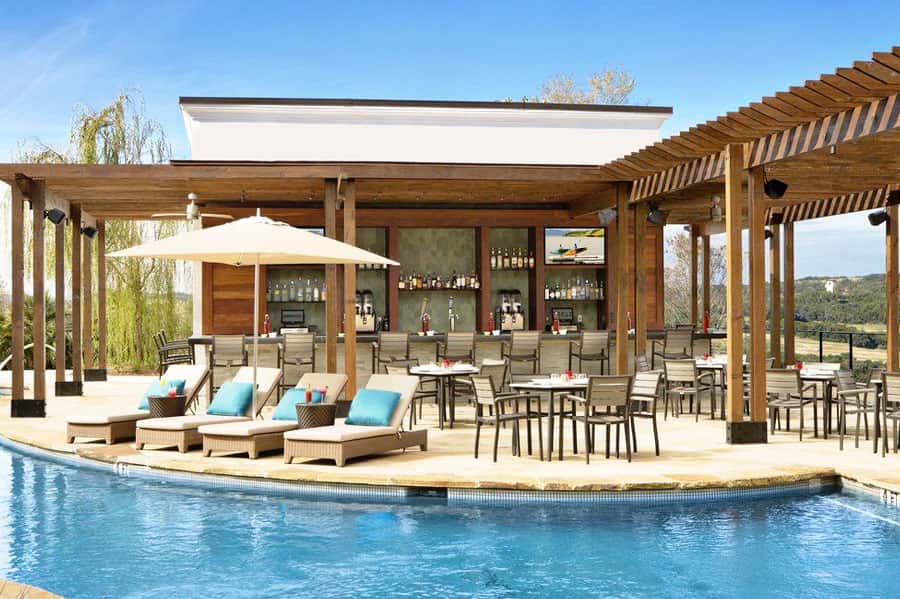 Luxury pool bar pavilion with wooden pergola, lounge chairs, dining area, and full-service bar overlooking resort grounds