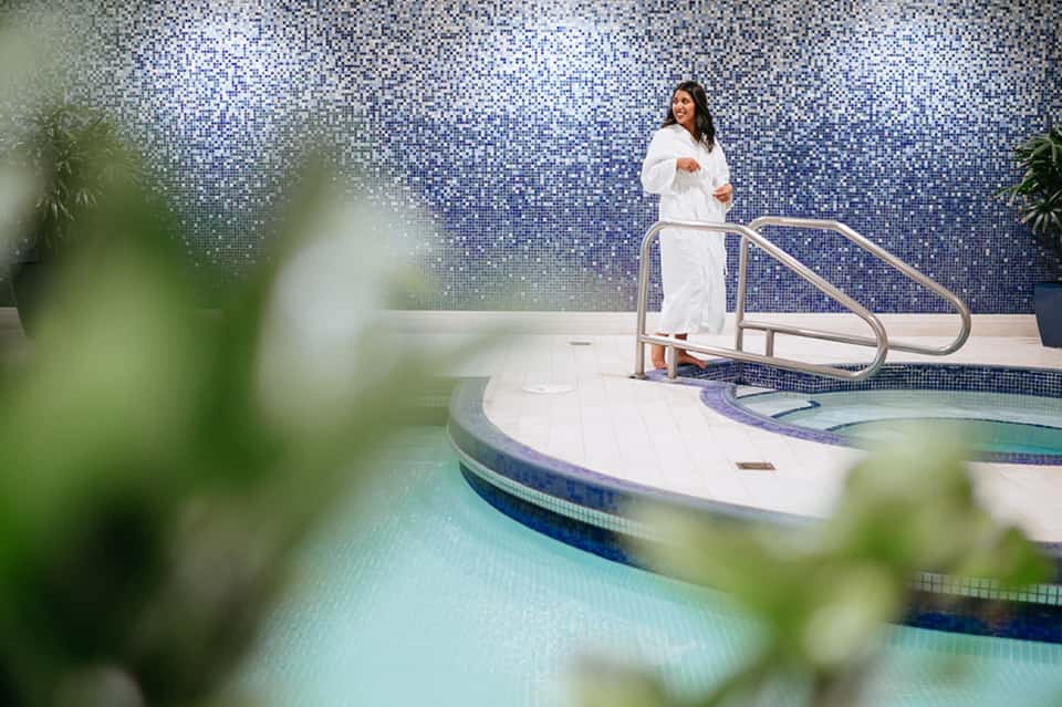 Woman in white robe standing by indoor spa pool with blue mosaic tile wall