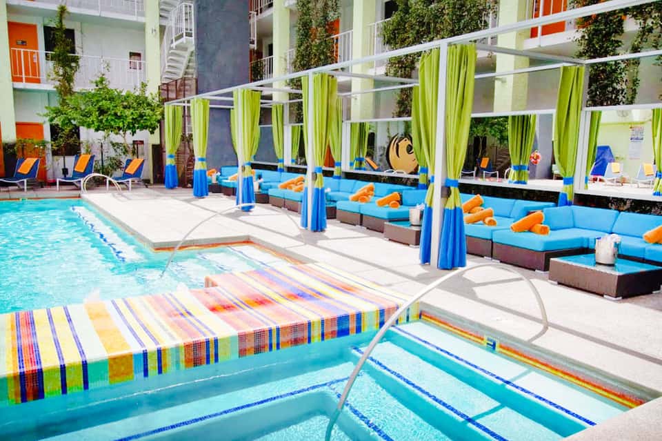 Resort pool courtyard with colorful lounge chairs, blue cabanas with green curtains, and lush green landscaping