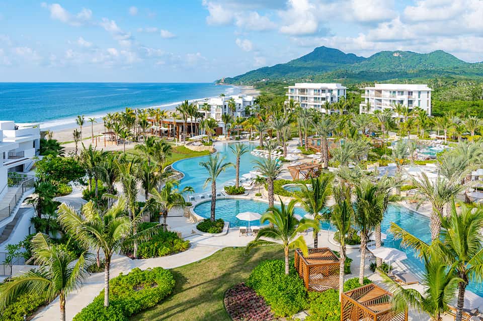 Aerial view of beachfront resort with pools, palm landscaping, and mountain backdrop