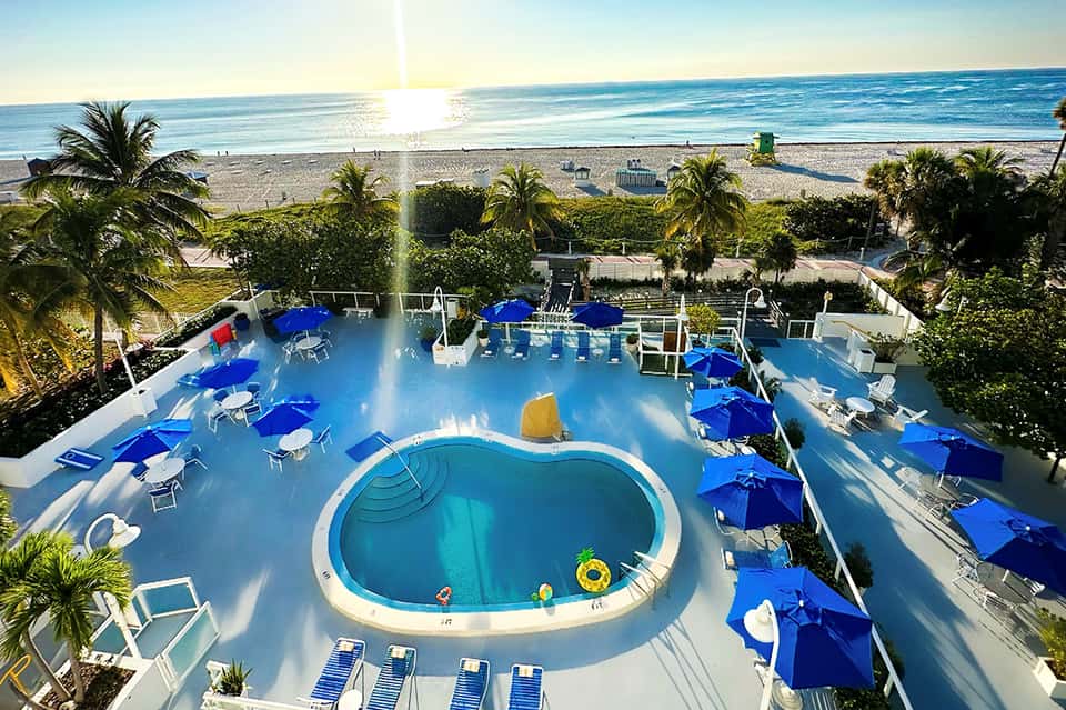 Aerial view of kidney-shaped pool with blue umbrellas overlooking ocean and beach
