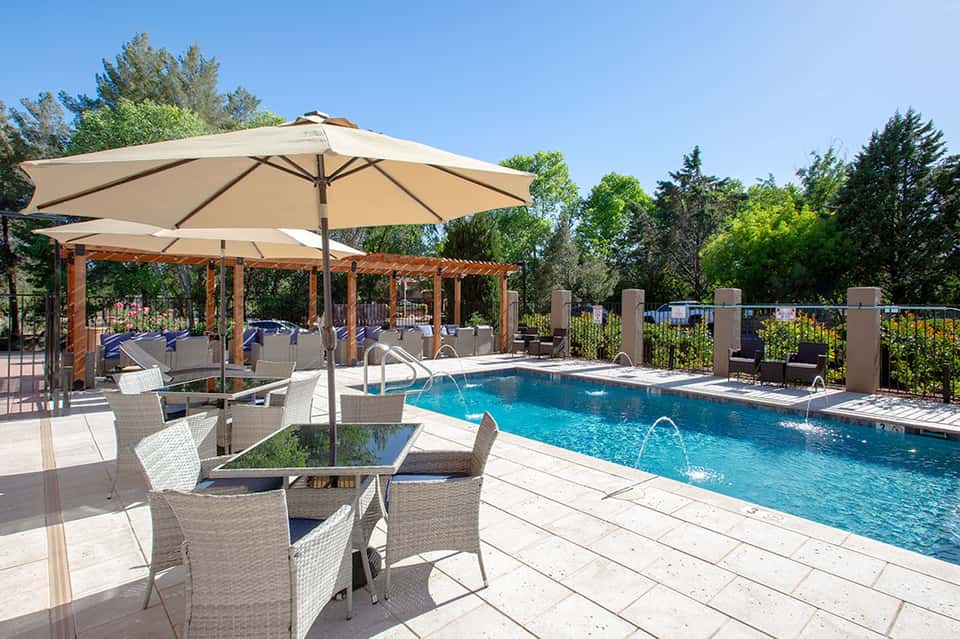 Resort pool area with turquoise water, large cream umbrella, wooden pergola, wicker lounge chairs, and green trees