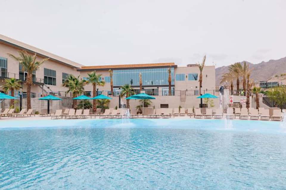 Resort pool with turquoise water, palm trees, blue umbrellas, and modern beige buildings