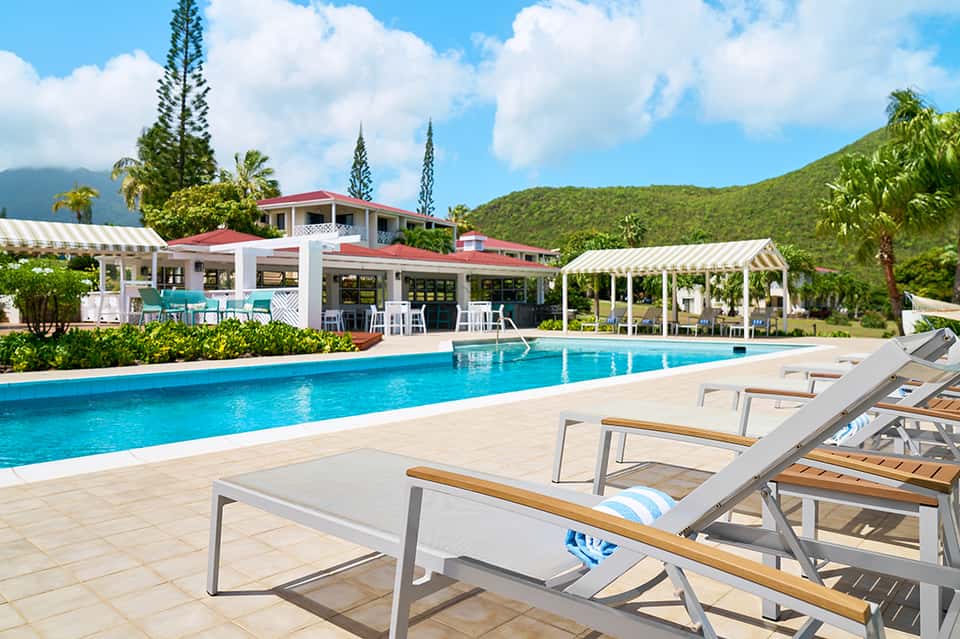 Resort pool with mountain backdrop, lounge chairs, cabanas with red roof, and tropical landscaping