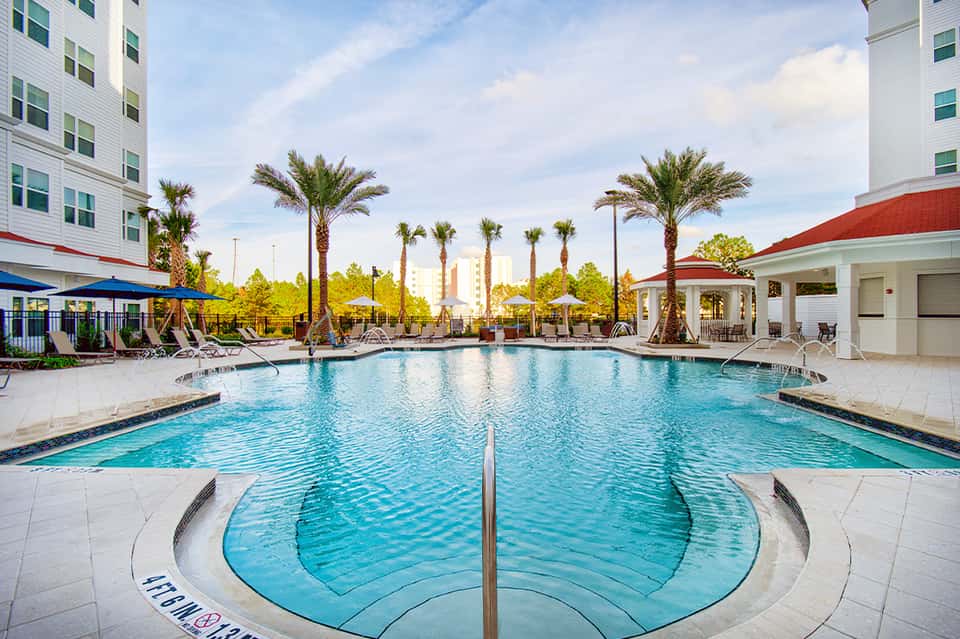 Resort outdoor pool surrounded by palm trees, white umbrellas, and modern hotel buildings