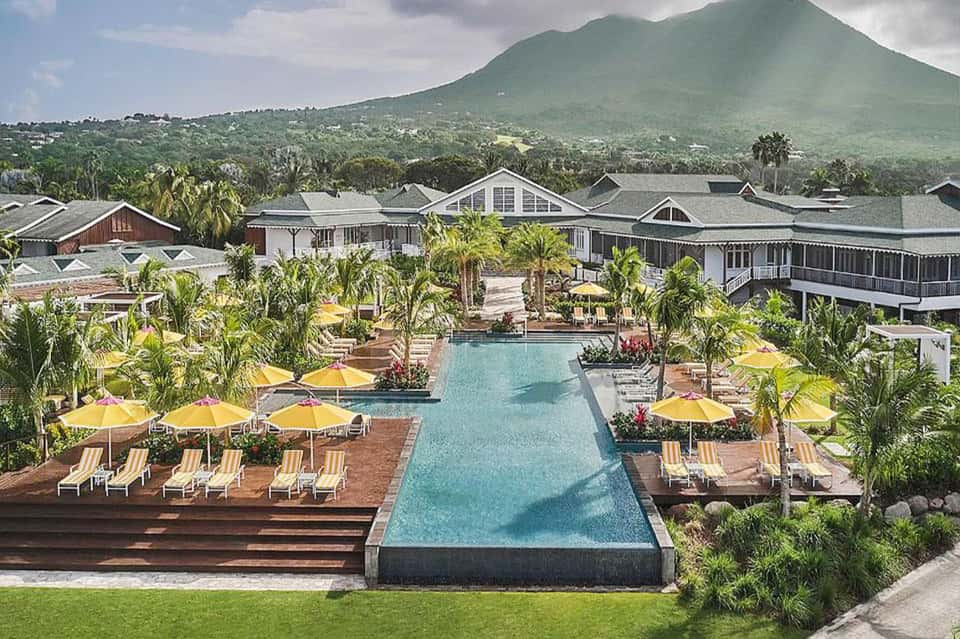 Aerial resort view with large pool, yellow umbrellas, tropical landscaping, and mountain backdrop