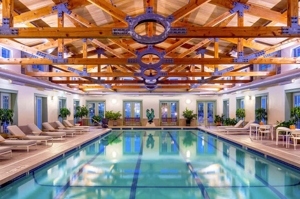 Indoor lap pool with decorative wooden beam ceiling, purple accents, loungers, plants, and blue water