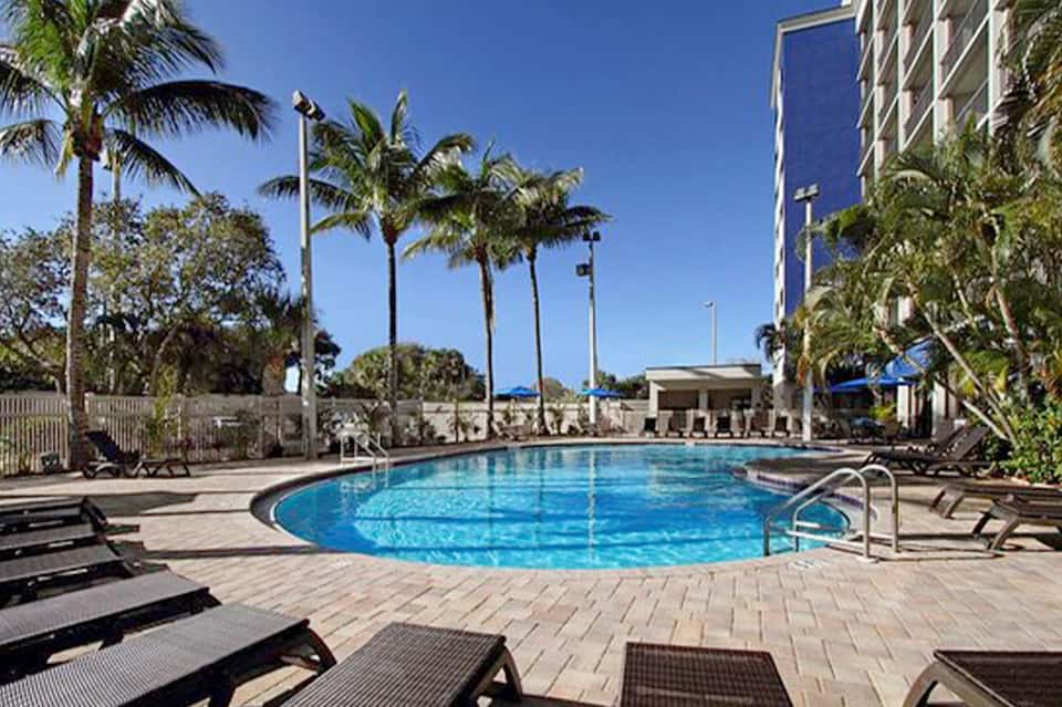 Circular blue pool surrounded by lounge chairs, palm trees, and tall residential building against clear sky