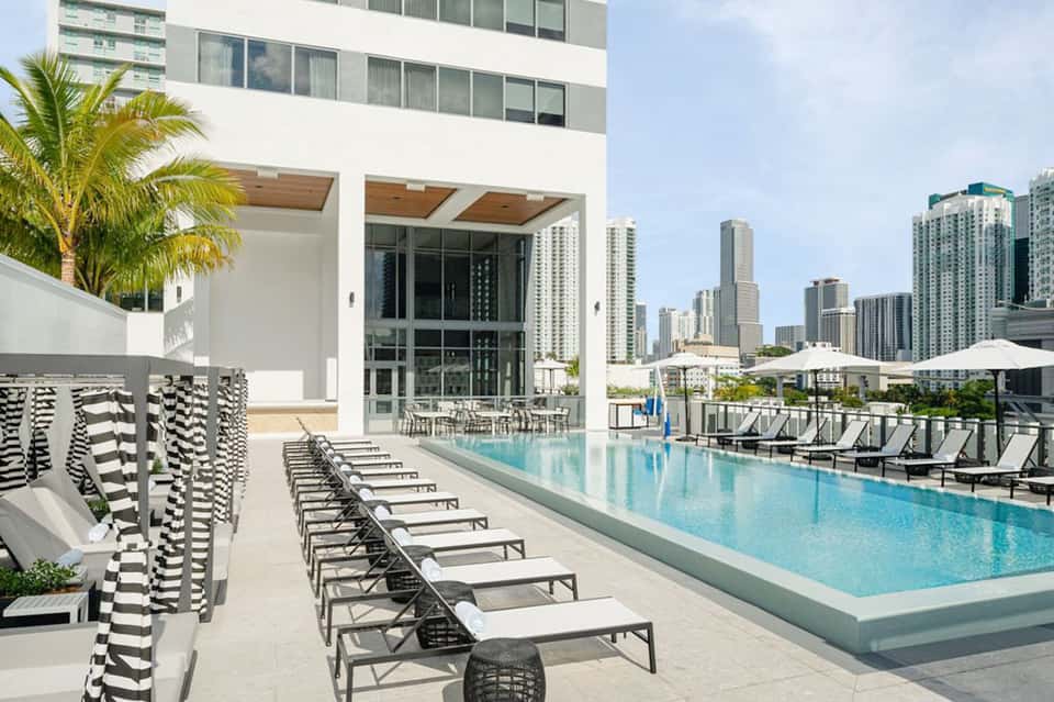 Modern rooftop pool with striped loungers, city skyline views, and white umbrellas in background