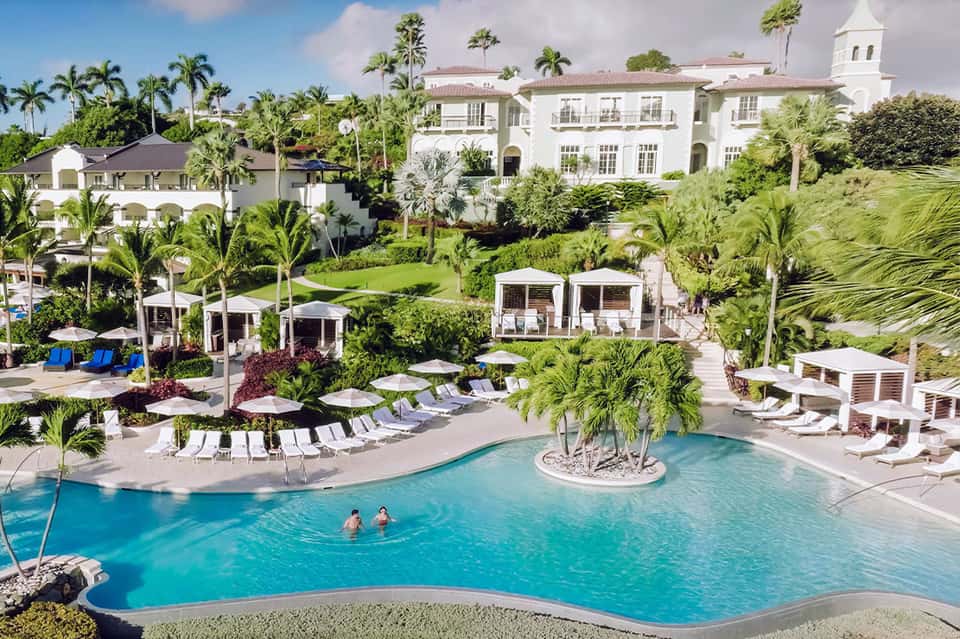 Luxury resort pool with island center, white cabanas, lounge chairs, palm trees, and colonial architecture