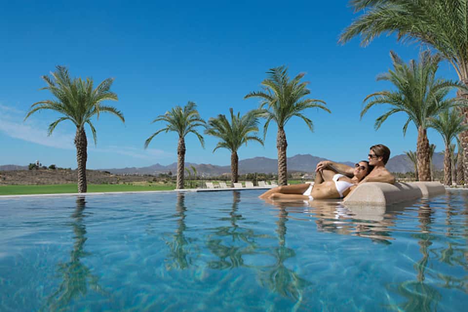 Two guests floating in infinity pool with palm trees and desert mountain landscape
