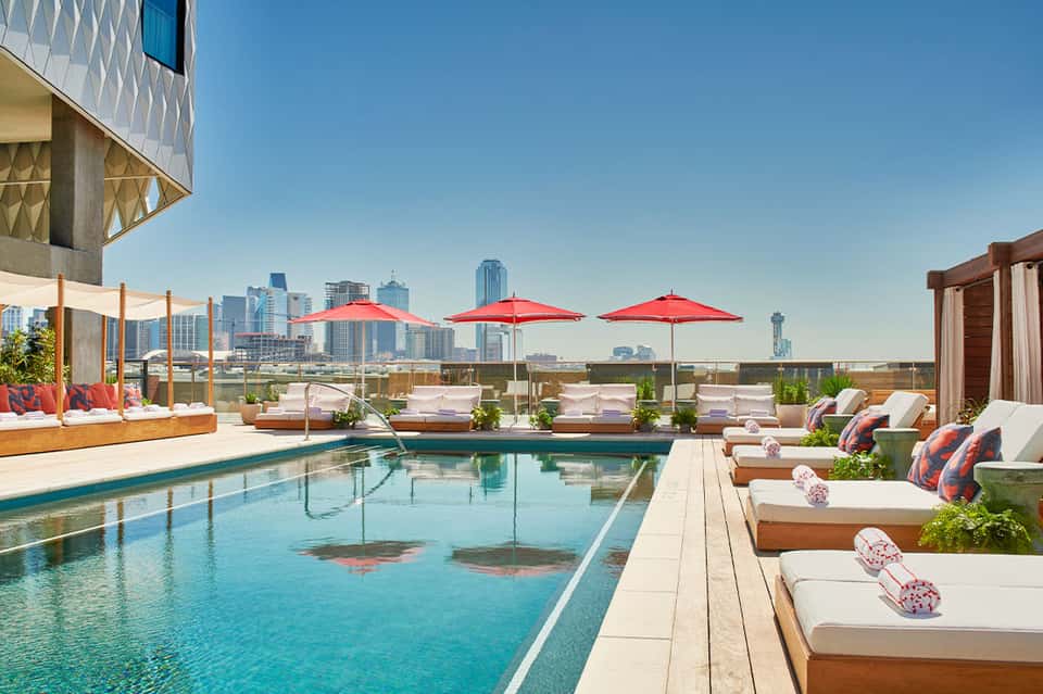 Urban rooftop pool with city skyline backdrop, red umbrellas, and lounge seating
