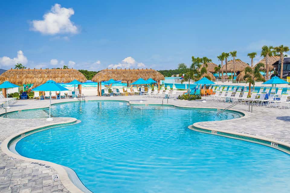 Resort pool with blue cabanas, thatched umbrellas, palm trees, and tropical landscape.