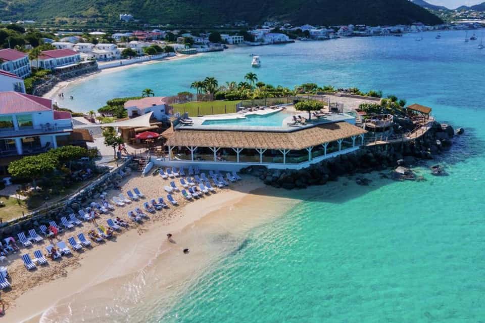 Aerial view of beachfront resort with pool, thatched pavilion, and crystal-clear turquoise waters