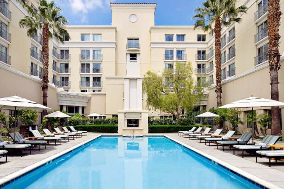 Resort courtyard pool with lounge chairs, umbrellas, and palm tree-lined buildings