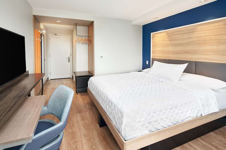 Compact hotel room with queen bed, blue accent wall, desk, and modern furnishings