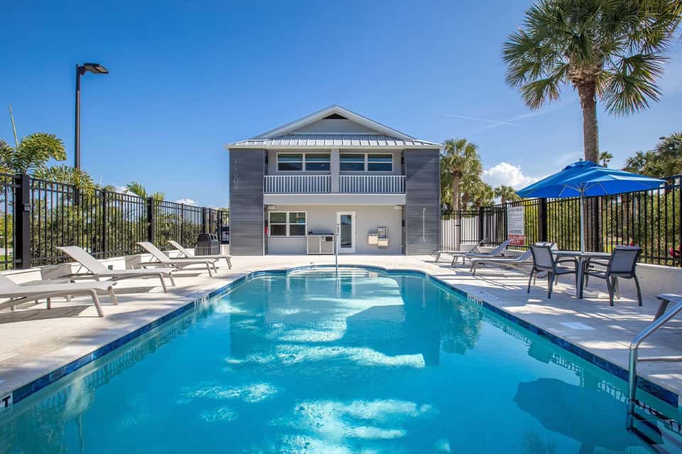 Rectangular lap pool with pool house, white lounge chairs, blue umbrellas, and palm trees