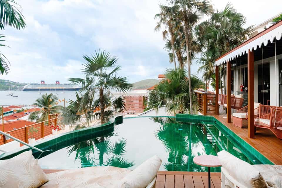 Oceanfront infinity pool overlooking cruise ship, palm trees, white loungers, wooden deck, and Caribbean view