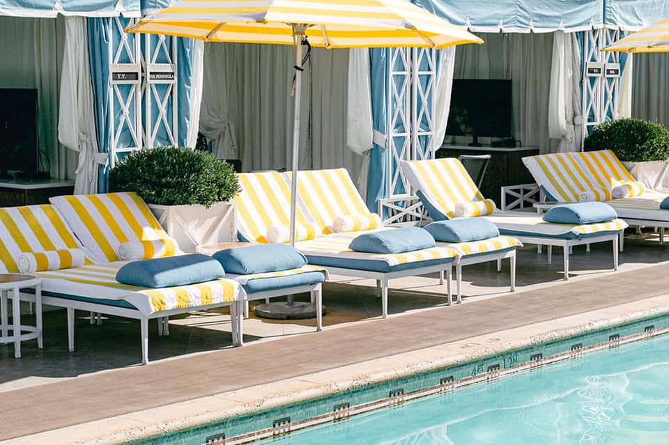 Pool cabanas with yellow and white striped umbrellas, teal curtains, lounge chairs, and calm pool water