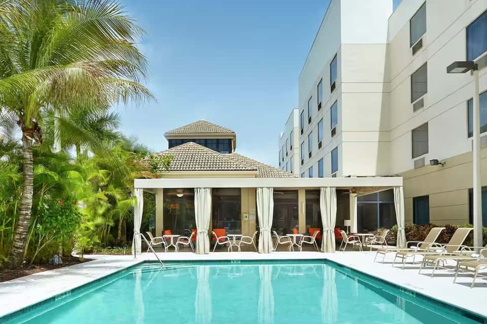 Hotel pool with white pergola cabana, loungers, palm trees, and modern building facade