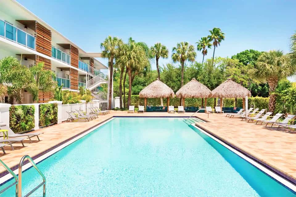 Resort pool with thatched umbrellas, lounge chairs, and modern building with palm trees under clear blue sky