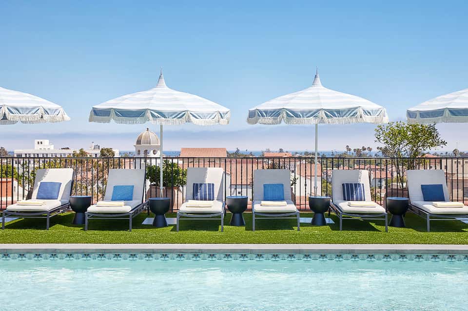 Poolside lounge area with white fringe umbrellas, white cushioned loungers, and city skyline view in background