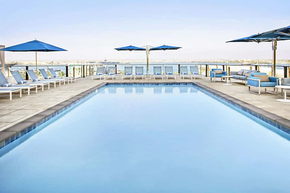 Rooftop infinity pool with blue umbrellas, lounge chairs, and city waterfront views