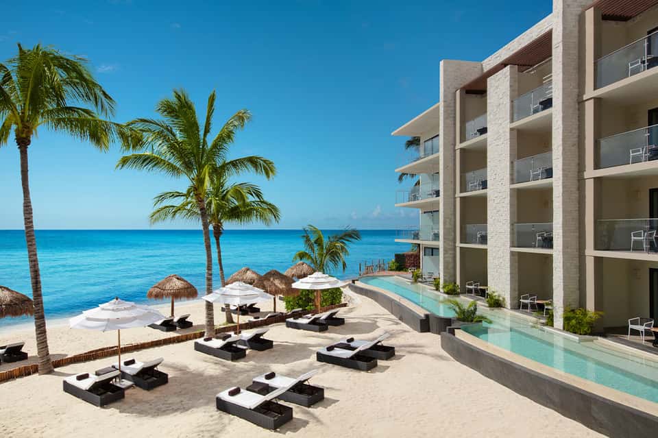 Beachfront resort with infinity pool, ocean views, black lounge chairs, palm trees, and modern concrete architecture