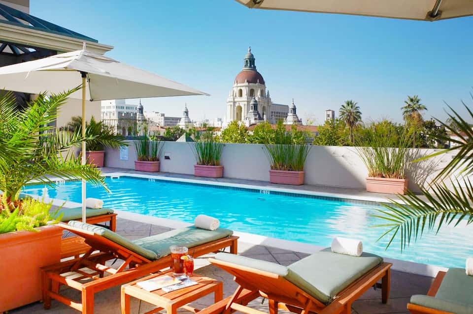 Rooftop pool with turquoise water, white loungers, palm plants, and cityscape with dome building in background