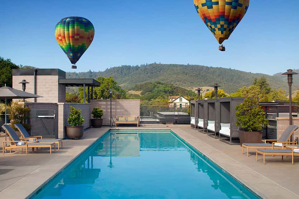 Rooftop pool with two hot air balloons floating overhead, mountains in background, loungers and modern architecture