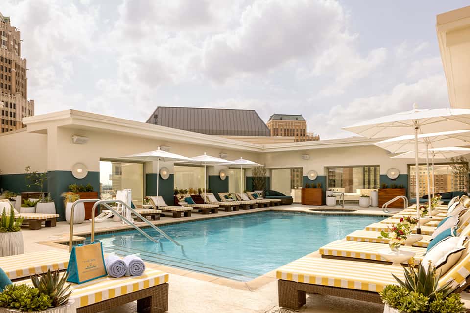 Urban rooftop pool with blue water, striped loungers, white umbrellas, and city skyline backdrop