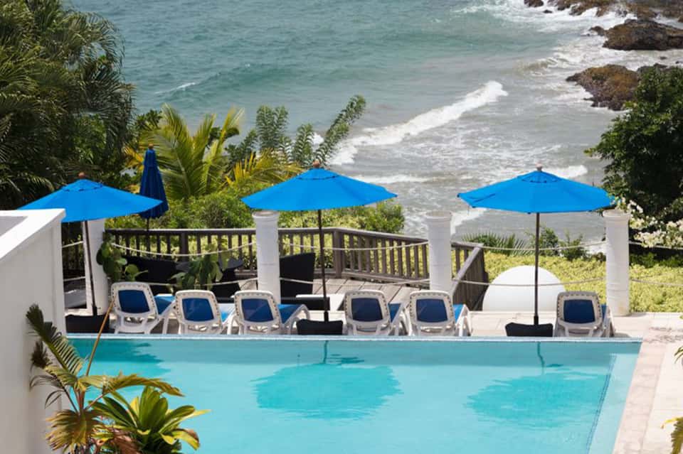 Oceanfront pool with blue umbrellas, lounge chairs, and ocean waves with rocky coastline