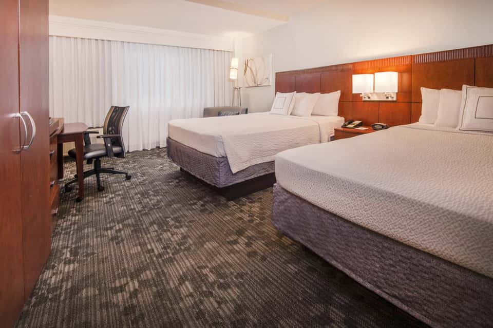 Hotel room with two queen beds, wood headboard, desk area, and white curtains with warm lighting