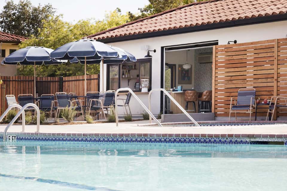 Poolside cabana with blue umbrella, outdoor seating, modern white building, and pool area