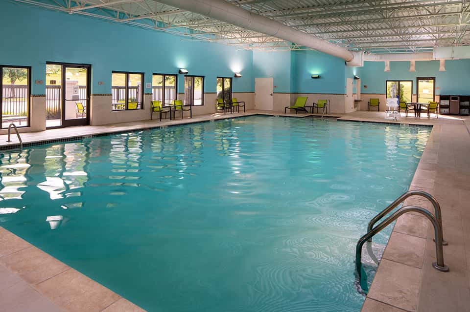 Indoor lap pool with turquoise water, bright walls, glass doors, and poolside seating