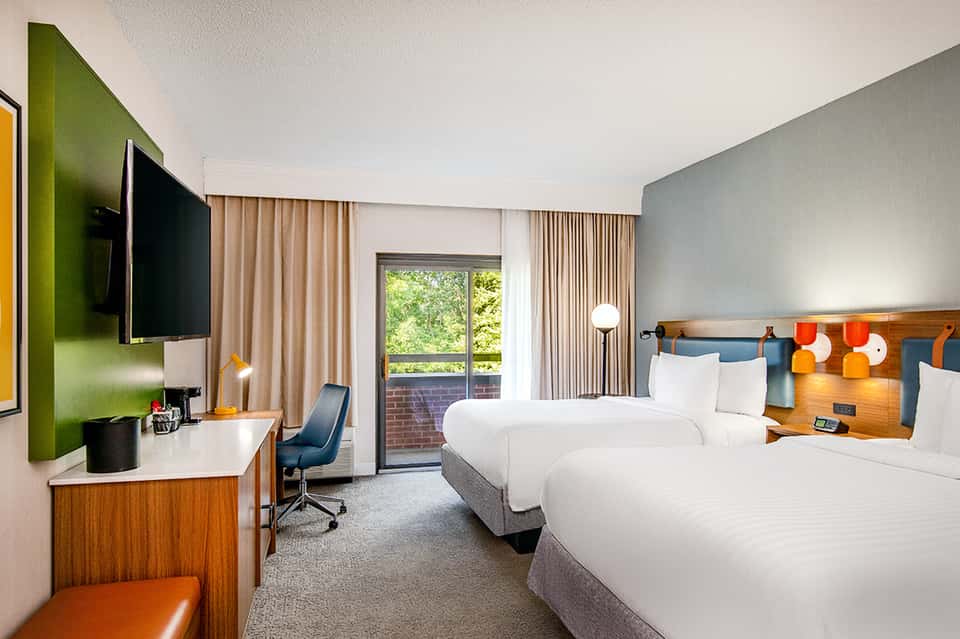 Contemporary hotel room with two queen beds, green accent wall, wood headboard, and balcony access