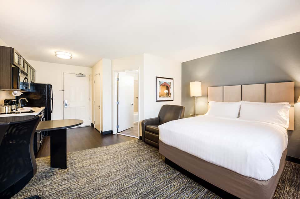 Hotel room with queen bed, work desk, kitchenette, and contemporary furnishings in neutral tones