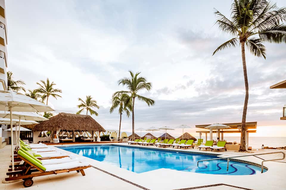 Beachfront resort pool at dusk with palm trees, white loungers, tiki hut, and ocean horizon
