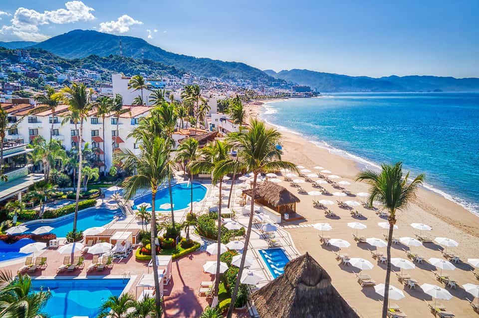 Beachfront resort with multiple pools, palm trees, sandy beach with loungers, and ocean mountain views