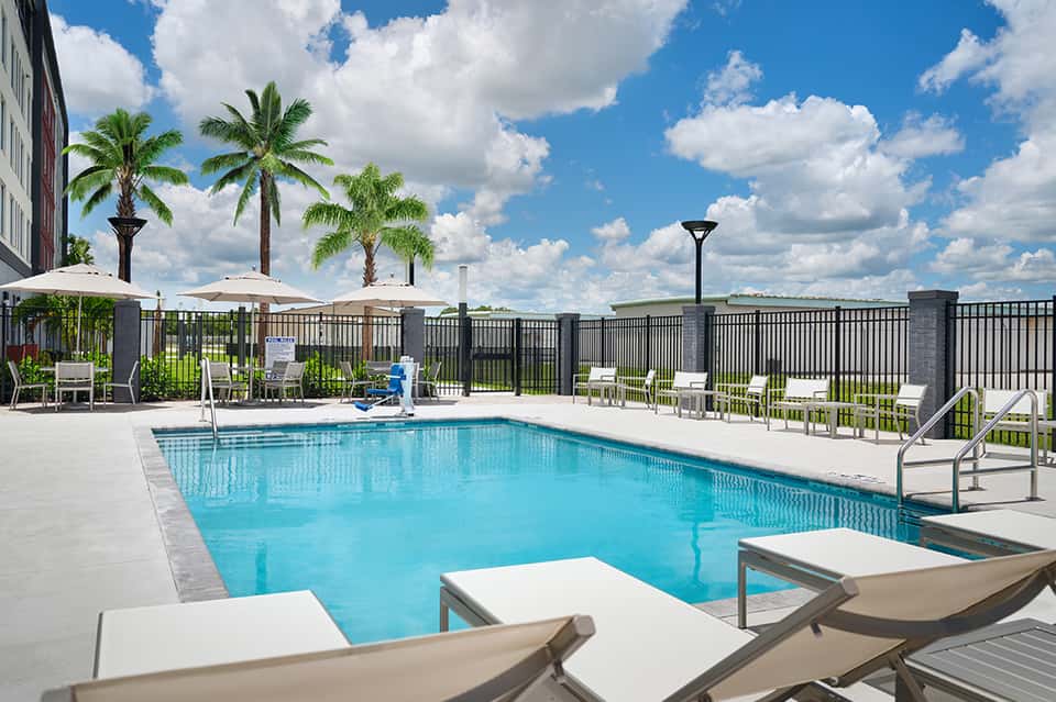 Resort pool with white lounge chairs, umbrellas, palm trees, and black fence railing