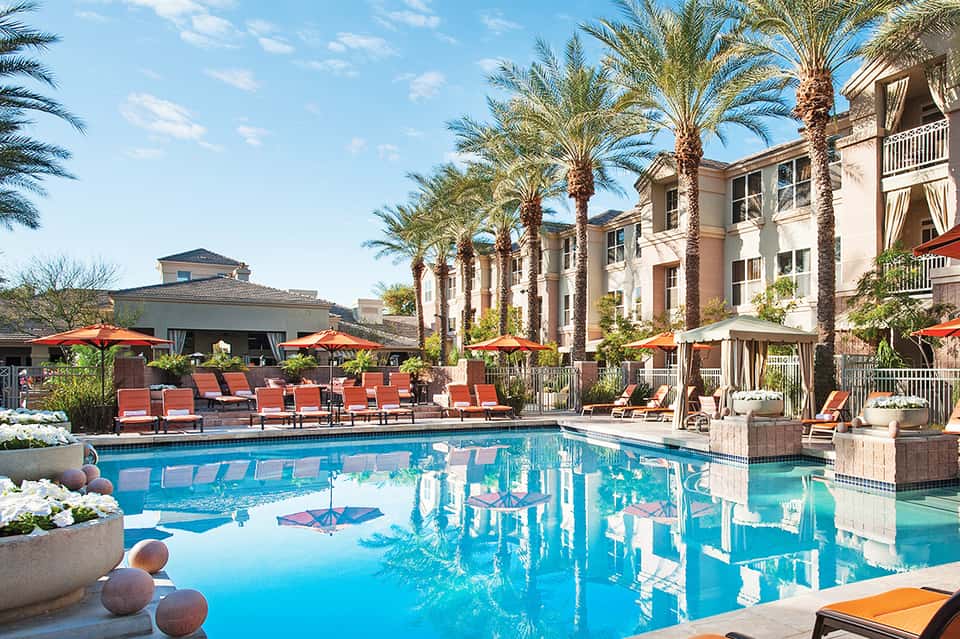 Resort pool area with blue water, red lounge chairs, palm trees, and building facade