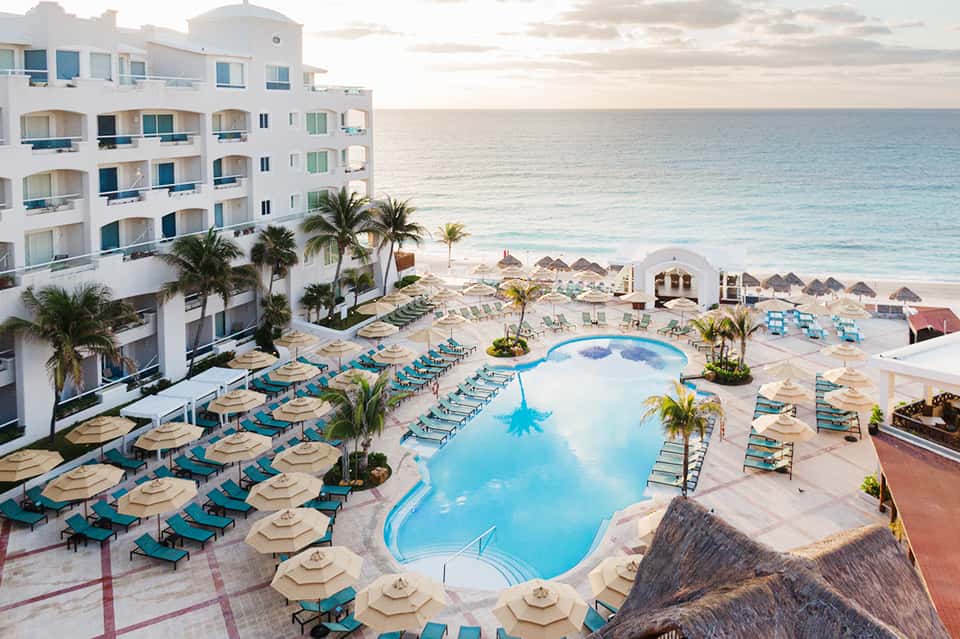 Beachfront resort pool with turquoise water, lounge chairs, palm trees, beach, ocean, and white hotel building