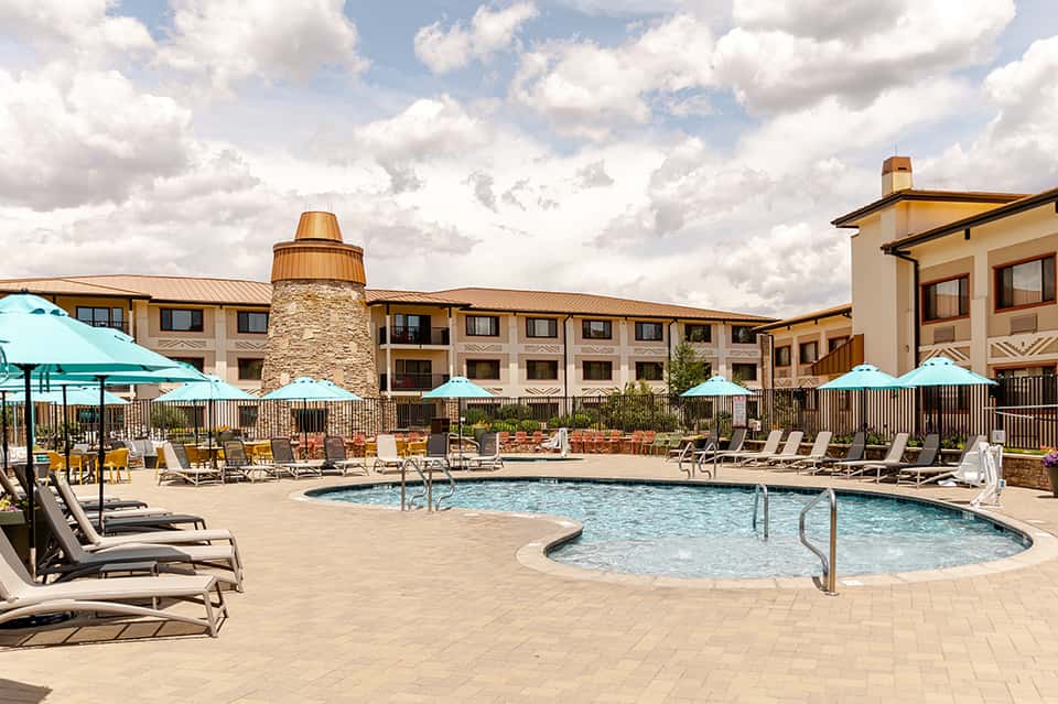 Resort pool area with loungers, umbrellas, and hot tub surrounded by stone and stucco buildings under cloudy sky